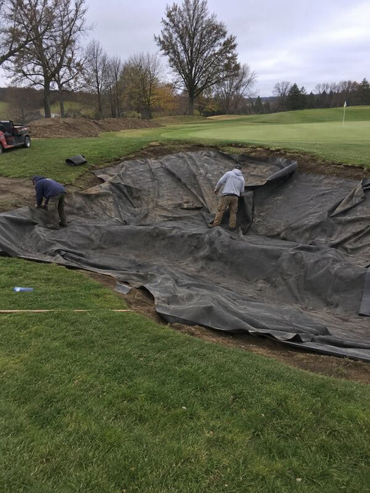 Crew installing bunker liner fabric in a golf course bunker near a lake with drainage pipe staged nearby
