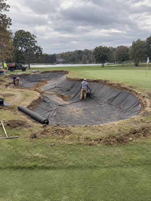 Rolled bunker liner fabric beside a bunker installation near a lakeside green

