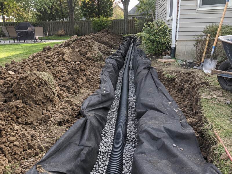 Non-woven geotextile fabric lining a French drain trench beside a backyard patio
