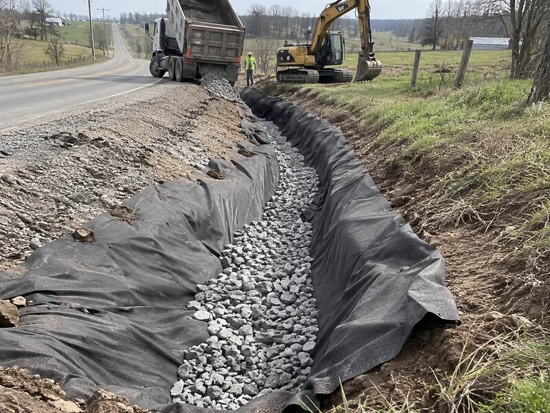 Dump truck delivering stone over non-woven geotextile fabric in a rural roadside drainage ditch
