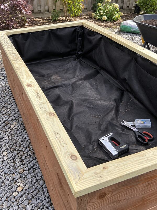 Staple gun and scissors resting on raised bed liner fabric inside a wood planter box
