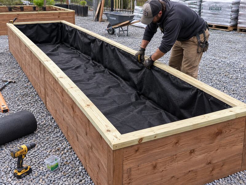 Worker installing non-woven raised garden bed liner fabric in a long wood planter box
