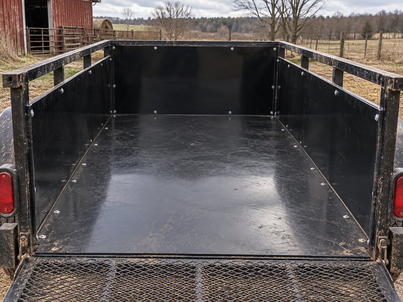Black HDPE trailer liner in a mesh-sided utility trailer parked near a farm barn and hay bales
